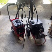 Two pressure washers side by side on concrete floor, one electric and red, the other gas-powered with a black tank.