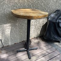 Full view of the bar-height table showing the wooden top and metal base on a deck next to a wall.