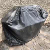 Barbecue with black fitted cover outdoors against a wall on paved surface.