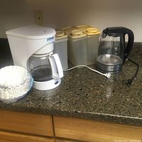Overall lot on kitchen counter showing white drip coffee maker with filter papers, three plastic storage containers with beige lids, and glass electric kettle.