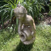 Front view of the weathered concrete cherub statue holding a large pitcher in a garden setting with green foliage background.