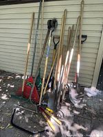 Various garden tools leaning against a wall including rakes, shovels, hoes, and a bow saw.