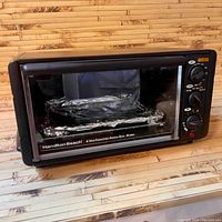 Front view of black Hamilton Beach toaster oven on wooden surface with aluminum foil tray inside.