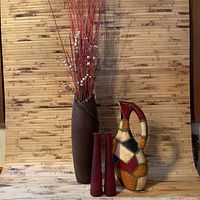 Home decor set showing a large brown vase with red branches and white berries, two slim red vases, and a colorful patchwork jug all placed on wooden surface with bamboo backdrop.