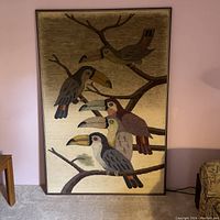 Front view showing entire framed textile art featuring five parrots on branches with cream background.