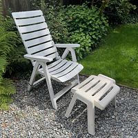 White lawn chair with slatted backrest and seat, shown outdoors on gravel and near garden plants