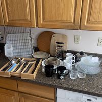 Overview of kitchen counter showing pottery bowls and plates, mugs, drinking glasses, wire dish rack, wooden cutting boards and kitchen utensils on countertop.