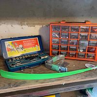 Photo showing the ratchet set in a blue case, orange compartment box with drawers, some metal hardware parts and a green plastic tool or handle on a wooden shelf.