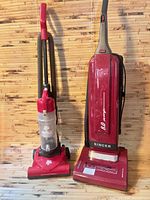 Two red upright vacuum cleaners side by side, one Singer labeled 8.0 amps, one Dirt Devil Quick Lite with transparent dust cup.