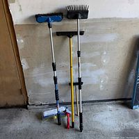 Four utility brushes leaning against a wall showing different sizes and handle types, including two with extendable poles.