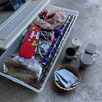 Full view of large clear storage container filled with assorted Christmas wrapping paper, gift bags and other decorative items. Set alongside five Christmas-themed mugs and a woven basket with cloth napkins.