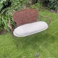 Photo showing the full bench on grass with decorative wrought iron frame, wicker backrest and beige cushion.