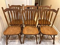 Front view showing all six solid oak wooden dining chairs arranged side by side, highlighting the contoured seats, vertical spindles on the backrests, and turned legs with stretchers.