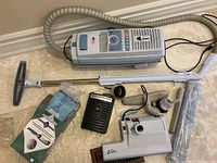 Full view of the Aerus Lux5500 vacuum cleaner, its flexible hose, metal extension wand, floor brush attachment, Electrolux filter bags, Electrolux 7000 accessory, and other cleaning heads laid out on a tiled floor.