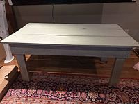 Front angled view of rustic painted solid wooden table showing top surface and legs on a patterned rug.