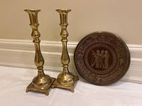 Two tall brass candlesticks and one round ornate brass plate together on floor against baseboard