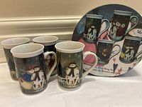 Four 12oz ceramic mugs with snowmen holiday images next to the original round decorative box showing mug images.