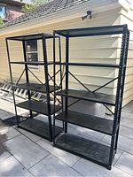 Two black metal garage/workroom shelves standing side by side outside, each with four shelves and metal cross bracing on the back for stability.