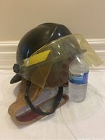 Black hard helmet with yellow face shield and tan ear flaps, standing upright next to a water bottle for scale.