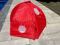 Photo of the red beach play tent closed and standing upright on patio near pool, showing front door and circular mesh window.