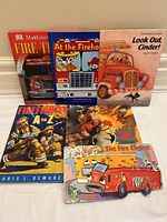 Set of six children's picture books with fire truck and firefighter themes, arranged standing and lying on the floor showing covers and attached toy fire truck.