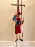 Full view of the Pinocchio marionette hanging by its strings showing the multicolored patchwork shirt, red pants, and controlling crossbar.
