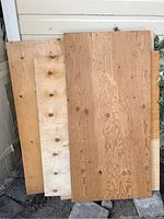 Five partial sheets of plywood of varying sizes and thickness leaning against a wall outside.