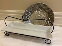 Photo of white ceramic rectangular serving dish on a black metal stand and round metal bowl with embossed vegetables.