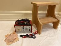 Photo showing battery charger and wooden step stool side by side, charger with cables and clips coiled on top, also instruction and warranty paper on floor