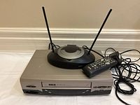 RCA VCR unit shown with TV antenna, remote control, and attached cables, photographed against wall baseboard.