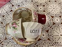 Four bone china cups and saucers stacked and arranged on a crocheted lace tablecloth, showing floral and gold designs.