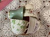 Four bone china cups and saucers stacked showing various patterns including floral, shamrocks, blue with gold trim, and red floral interior.