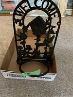 Front view of black cast iron plant hanger positioned inside a cardboard box, showing the welcome arch and birdhouse design.