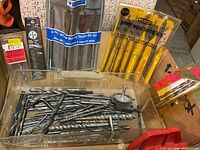 Photo showing assortment of drill bits including boxed wood boring bits, spade bits in packaging, and various loose metal bits along with wire brush and grinding wheel attachments.