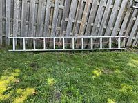 Full view of ladder in a retracted position lying on grass with wooden fence background
