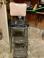 Full view of kitchen stool showing padded beige vinyl backrest, black vinyl seat with tear and paint splatters, and metal frame with two front steps.