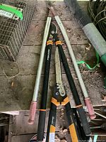 Top down view of three pruning tools placed on a wooden surface, showing Fiskars pruner centered with two long-handled metal loppers on each side.