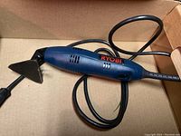 Ryobi detail sander in blue and black with power cord coiled around, triangular sanding pad attached inside a cardboard box.