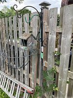 Black metal shepherd's hook hanging on a wooden fence, featuring a round metal basket attached under a curled hook.