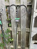 Two black metal shepherd’s hooks leaning against a wooden fence, showing both hooks and spiral decorative details at the top.