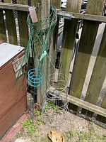 Green, teal, and silver metal tomato cages and plant supports leaning against wooden fence outdoors, showing multiple types of wireframe support structures for plants.