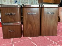 Four vintage woodgrain canisters labeled coffee, tea, sugar, flour, shown standing on a pink tablecloth.