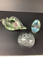 Three glass paperweights displayed on a black surface: sea shell shaped paperweight, signed iridescent oval paperweight, and clear round snowflake paperweight.
