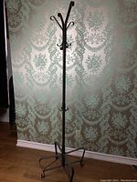 Full view of the metal coat tree stand against a patterned wallpaper background, showing its height and tripod base.