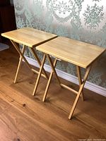 Pair of wooden TV tables with rectangular tops and foldable legs shown standing open on hardwood floor against patterned wallpaper.