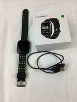 Smart watch with strap, original box featuring watch image and digital time, and USB charging cable arranged on white cloth background