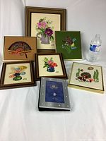 Overview of all items in the lot: six framed small needlepoint artworks featuring floral and nature motifs, plus one silver-tone photo frame. Water bottle shown for scale.