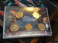 Front view of 5 gold plated Barack Obama quarters in a display case with images of Obama and gold plated coins, case cracked at bottom right corner.