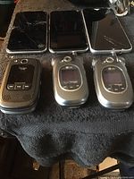 Top-down view of six cell phones arranged in two rows on dark fabric background showing cracked screens on smartphones and external features of flip phones.