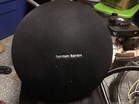 Front view of black Harman Kardon round speaker with logo visible on fabric grille.